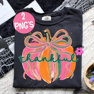 Peut inclure: T-shirt noir avec un motif de citrouille color&eacute;e. La citrouille est orange, rose et dor&eacute;e, avec un n&oelig;ud vert et rose. Le mot "thankful" est &eacute;crit en vert. Un cercle rose avec "2 PNG'S" est visible.