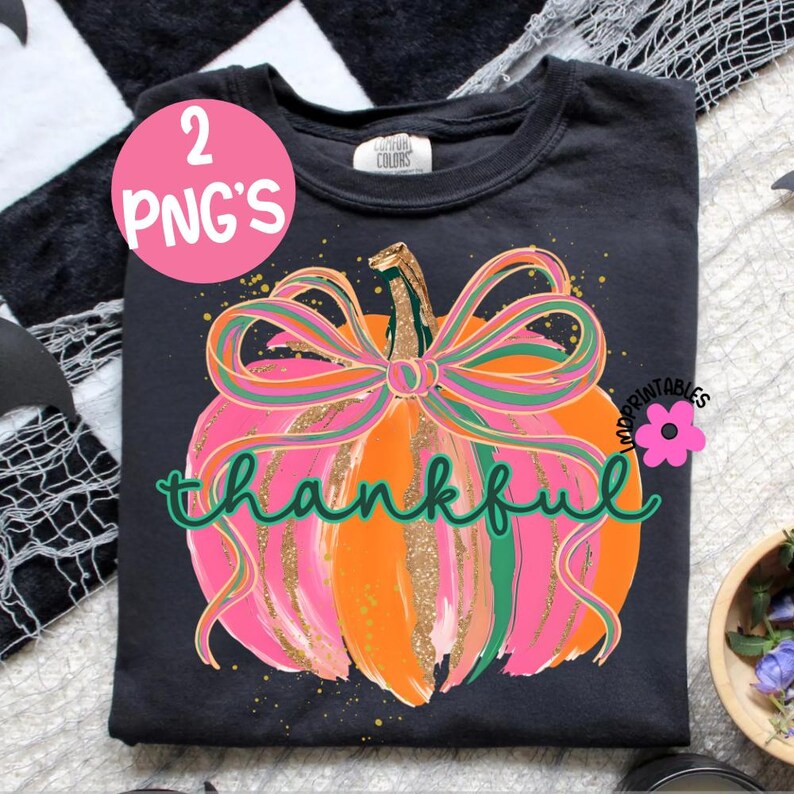 Peut inclure: T-shirt noir avec un motif de citrouille color&eacute;e. La citrouille est orange, rose et dor&eacute;e, avec un n&oelig;ud vert et rose. Le mot "thankful" est &eacute;crit en vert. Un cercle rose avec "2 PNG'S" est visible.