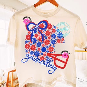 May include: Cream-colored t-shirt featuring a football helmet design made of red and blue flowers, with a blue bow and the word "gameday" in script. The shirt also has the text "original" in a circle.