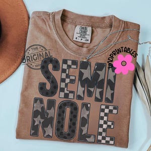 May include: A brown t-shirt with the word "SEMINOLE" in a mix of patterns, including stars, polka dots, and checkered designs. The shirt has a crew neck and short sleeves. A silver necklace is draped across the shirt.