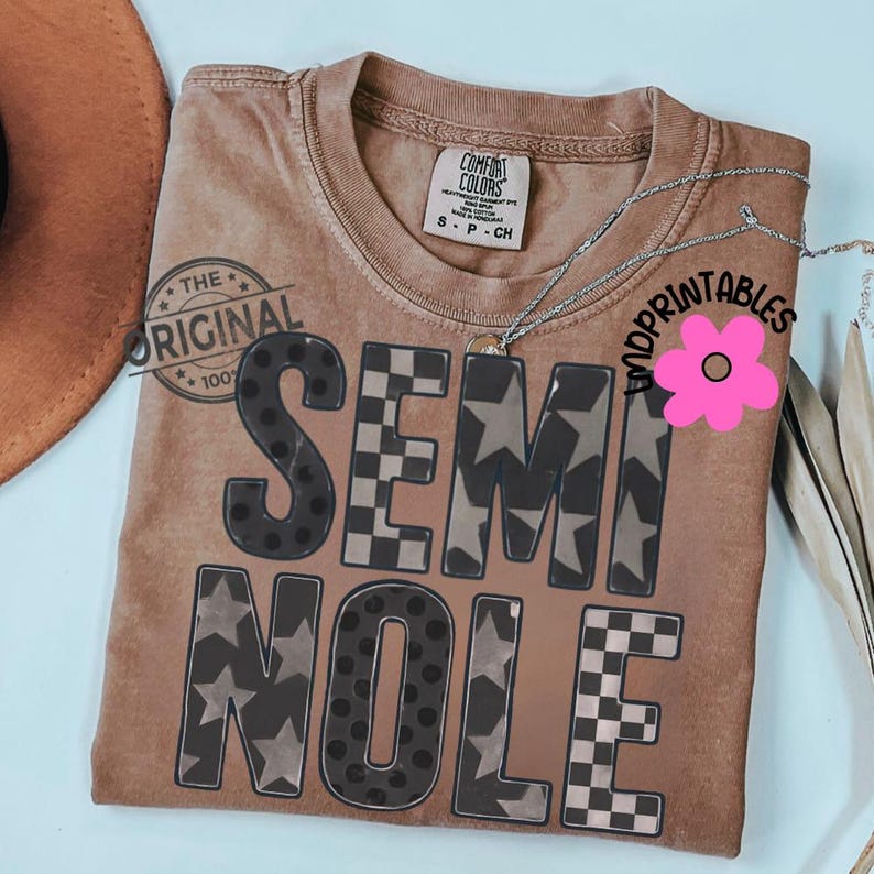 May include: A brown t-shirt with the word "SEMINOLE" in a mix of patterns, including stars, polka dots, and checkered designs. The shirt has a crew neck and short sleeves. A silver necklace is draped across the shirt.