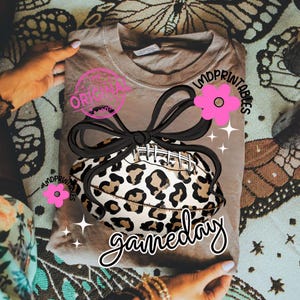 May include: A tan t-shirt featuring a football with a leopard print design, a black bow, and pink flower accents. The shirt has the text "gameday" in a script font, and the word "original" in a pink stamp.