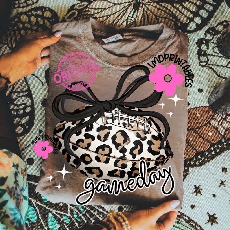 May include: A tan t-shirt featuring a football with a leopard print design, a black bow, and pink flower accents. The shirt has the text "gameday" in a script font, and the word "original" in a pink stamp.