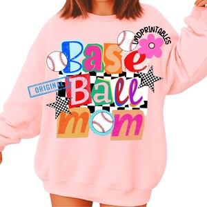 以下が含まれることがあります： 明るいピンク色のスウェットシャツ。カラフルなブロックレターで「Base Ball Mom」と書かれています。デザインには野球ボール、花、市松模様の星が含まれています。「ORIGINAL」の文字が左側にスタンプされています。