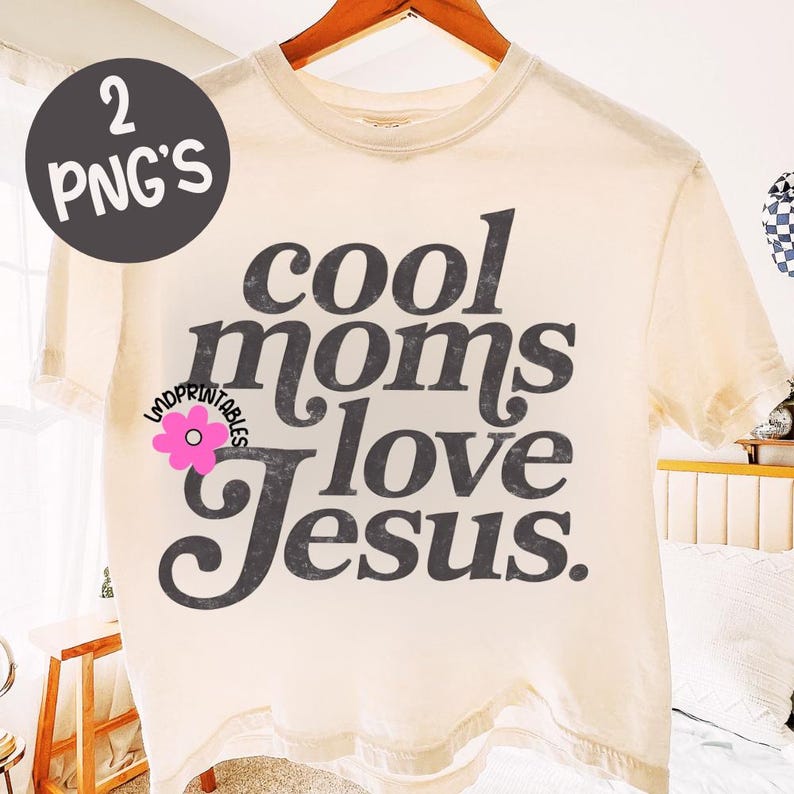 Puede incluir: Camiseta color crema con el texto "cool moms love Jesus" en gris oscuro, con un estilo vintage. Un gr&aacute;fico de una flor rosa est&aacute; en el lado izquierdo de la camiseta. La camiseta se muestra en una percha de madera.