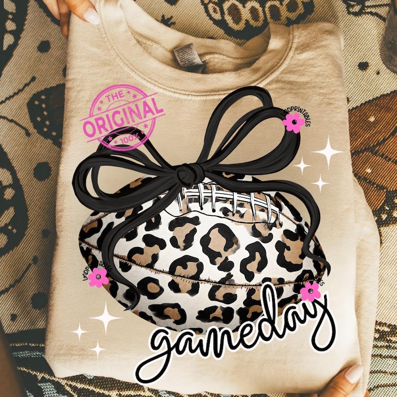 May include: A beige sweatshirt featuring a football with a leopard print design, tied with a black bow. The word "gameday" is written in a script font. The text "The Original" is stamped on the upper left.