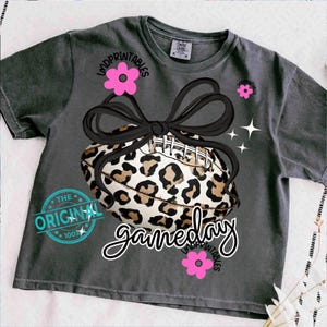 May include: Dark gray t-shirt featuring a leopard print football tied with a black bow. The shirt has the text "gameday" in a script font, along with pink flower graphics and the text "LMDPRINTABLES". The shirt also has the text "THE ORIGINAL 100%" in a blue circle.