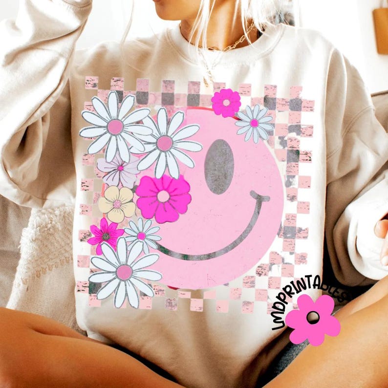Op de afbeelding: Cr&egrave;mekleurige sweatshirt met een grote roze smiley en bloemenontwerp. De smiley wordt omringd door witte, roze en gele madeliefjes. De achtergrond heeft een roze en grijs geblokt patroon.