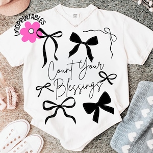 K&ouml;nnte beinhalten: Wei&szlig;es T-Shirt mit schwarzen Schleifengrafiken und dem Schriftzug "Count Your Blessings". Das Shirt hat einen V-Ausschnitt und kurze &Auml;rmel. Eine rosa Blumengrafik mit dem Wort "Imprintables" befindet sich oben links.
