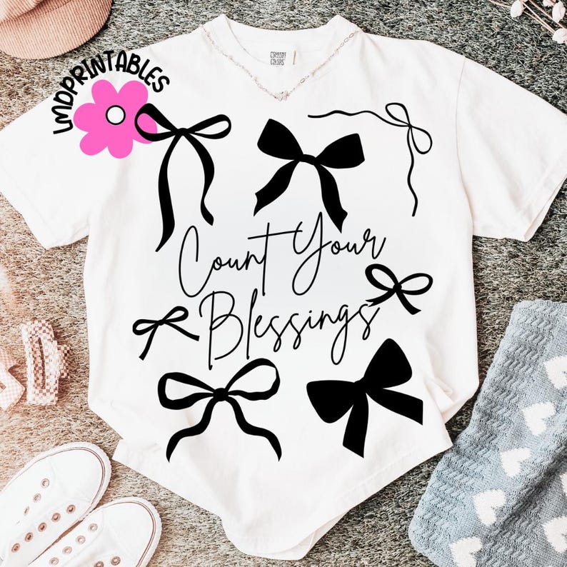 K&ouml;nnte beinhalten: Wei&szlig;es T-Shirt mit schwarzen Schleifengrafiken und dem Schriftzug "Count Your Blessings". Das Shirt hat einen V-Ausschnitt und kurze &Auml;rmel. Eine rosa Blumengrafik mit dem Wort "Imprintables" befindet sich oben links.