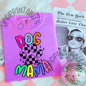 May include: A lilac-colored t-shirt with the words "DOG MAMA" in a colorful, playful font. The design includes a paw print with a checkered pattern. A newspaper and a disco ball are in the background.