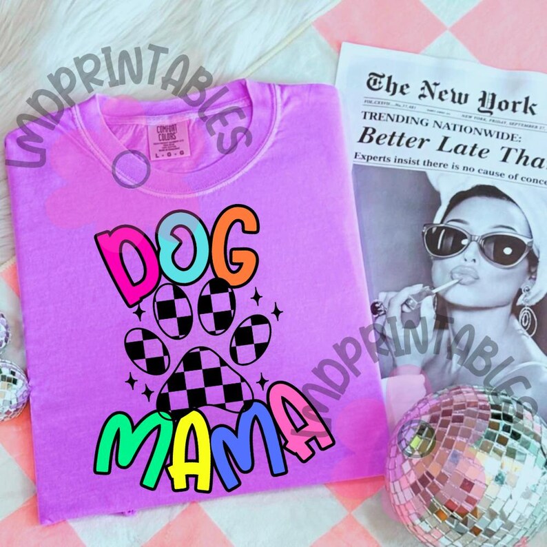 May include: A lilac-colored t-shirt with the words "DOG MAMA" in a colorful, playful font. The design includes a paw print with a checkered pattern. A newspaper and a disco ball are in the background.