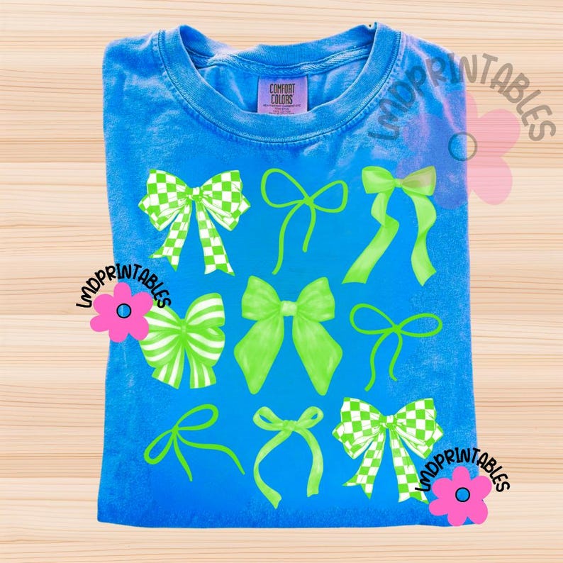 May include: A blue t-shirt with a variety of green bows. Some bows have a checkered pattern, while others are solid green. The shirt has the Comfort Colors brand tag. The text "LMDPrintables" is on the shirt.