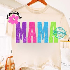 Pu&ograve; includere: T-shirt color crema con la scritta "MAMA" a grandi lettere colorate e invecchiate. Le lettere sono viola, rosa, blu e verdi. Sono visibili anche un logo floreale e un timbro circolare con la scritta "The Original".
