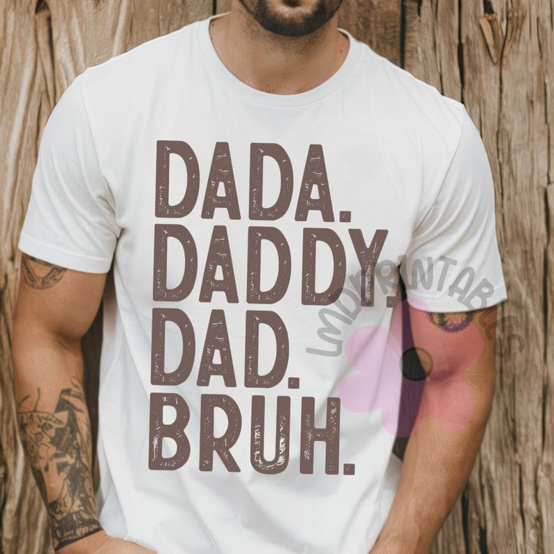 May include: White t-shirt with the words "DADA. DADDY. DAD. BRUH." printed in a brown, distressed font. The shirt is a crew neck style and appears to be made of a soft material. The background is a wooden surface.