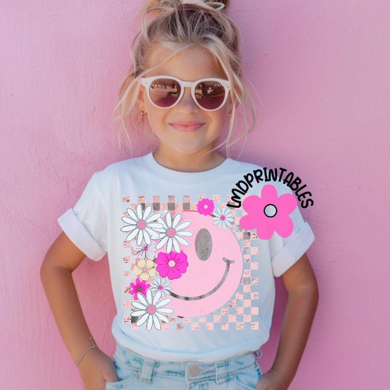 Op de afbeelding: Wit T-shirt met een roze smiley-gezicht, versierd met madeliefjes en bloemen. De smiley staat tegen een geruite achtergrond. De tekst "LMDPRINTABLES" is op het shirt gedrukt. Het meisje draagt een zonnebril.
