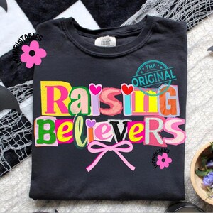 May include: Dark gray t-shirt with the words "Raising Believers" in a colorful, cut-out style. The design includes hearts and a pink bow, with the text "The Original" in a circular stamp.