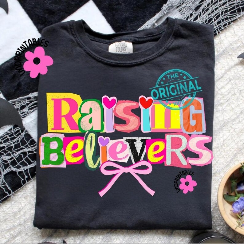 May include: Dark gray t-shirt with the words "Raising Believers" in a colorful, cut-out style. The design includes hearts and a pink bow, with the text "The Original" in a circular stamp.
