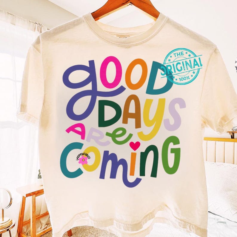 Może przedstawiać: Kremowa koszulka z napisem "Good Days Are Coming" w kolorowej, zabawnej czcionce. Projekt zawiera serce i grafikę kwiatową. Fraza "The Original 100%" jest wydrukowana w prawym g&oacute;rnym rogu.