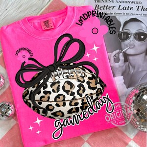 May include: A bright pink sweatshirt featuring a football with a leopard print design and a black bow. The word "gameday" is written in a cursive font. The text "LMDPRINTABLES" is also visible.