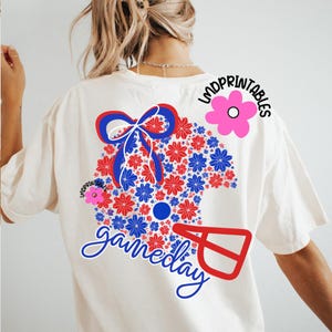 May include: Cream-colored t-shirt with a floral design in red and blue, shaped like a helmet. A blue bow and the word "gameday" in script are featured. The text "LMDPRINTABLES" is also visible.