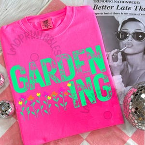 May include: A bright pink t-shirt with the word "GARDENING" in large, distressed green letters. Below the text is a row of colorful flowers. The shirt is a Comfort Colors brand.