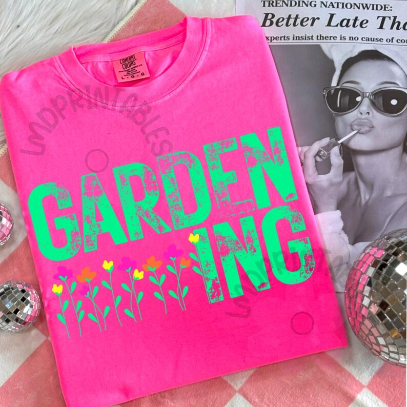 May include: A bright pink t-shirt with the word "GARDENING" in large, distressed green letters. Below the text is a row of colorful flowers. The shirt is a Comfort Colors brand.