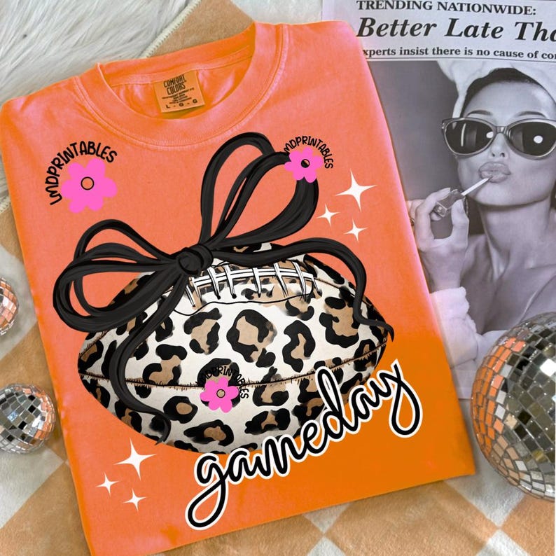 May include: Orange Comfort Colors sweatshirt with a leopard print football design, black bow, and the word "gameday". Pink flower accents and white starbursts complete the design. The text "MDPRINTABLES" is also visible.