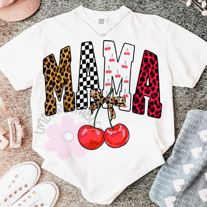 May include: White t-shirt with the word "MAMA" in a mix of leopard print, checkered, and cherry patterns. A bow and two red cherries are below the word. The shirt is a casual style.