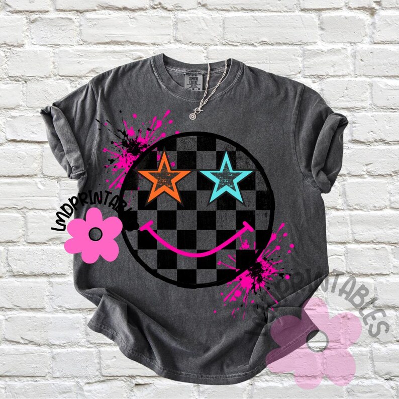 May include: Dark grey t-shirt with a smiley face design. The design features star-shaped eyes in orange and blue, a checkered pattern, and pink paint splatters. A silver necklace is visible. The shirt has rolled-up sleeves.