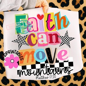 May include: White t-shirt with a colorful graphic design. The design features the words "Faith Can Move Mountains" in various fonts and colors, along with a pink flower and two checkered stars. The text "Matthew 17:20" is also included.