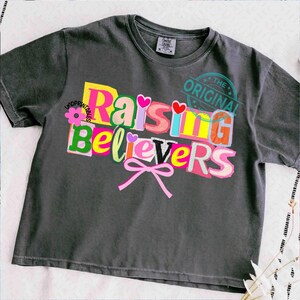 May include: Dark gray t-shirt with the text "Raising Believers" in a colorful, cut-out style design. The design includes hearts, a flower, and a pink bow. The words "The Original" are stamped on the upper right.