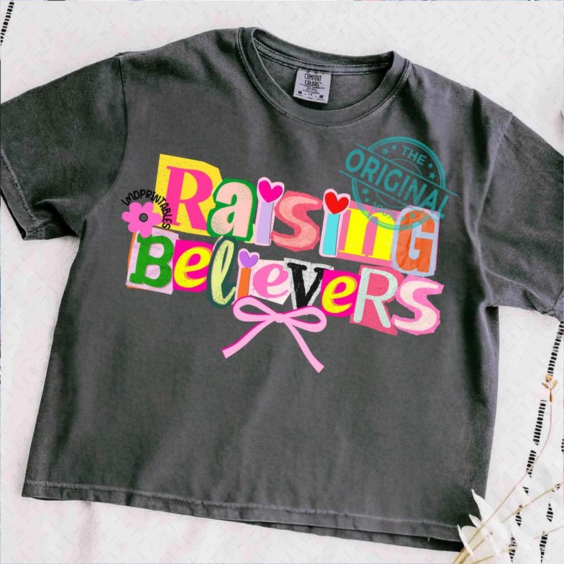 May include: Dark gray t-shirt with the text "Raising Believers" in a colorful, cut-out style design. The design includes hearts, a flower, and a pink bow. The words "The Original" are stamped on the upper right.