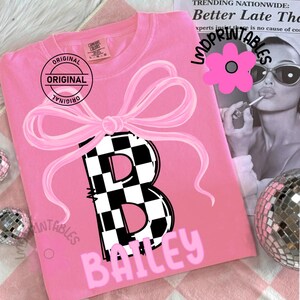May include: Pink Comfort Colors t-shirt with a large pink bow graphic and a black and white checkered letter "B". The name "BAILEY" is printed in pink below the letter. The shirt has the word "ORIGINAL" printed on it.