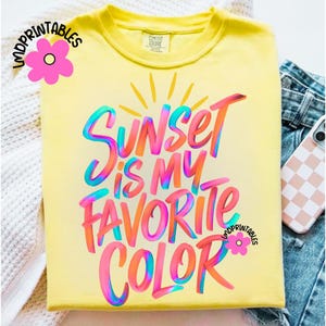 May include: A yellow t-shirt with the phrase "Sunset is my Favorite Color" in colorful, stylized lettering. The design includes a sun graphic and is from "LMDPrintables". The shirt is folded, with a phone case and denim visible.