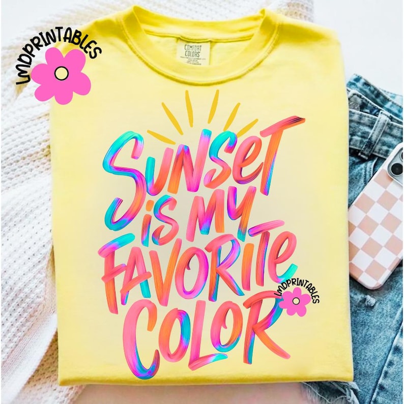 May include: A yellow t-shirt with the phrase "Sunset is my Favorite Color" in colorful, stylized lettering. The design includes a sun graphic and is from "LMDPrintables". The shirt is folded, with a phone case and denim visible.