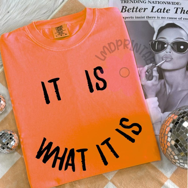 May include: Bright orange Comfort Colors t-shirt with the phrase "IT IS WHAT IT IS" in black lettering. The shirt is laid flat, showcasing the design. A newspaper clipping and disco ball are in the background.