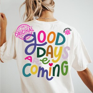 Może przedstawiać: Kremowa koszulka z napisem "Good Days Are Coming" w kolorowych, zabawnych literach. Projekt zawiera r&oacute;żową grafikę kwiatową i okrągłą pieczęć z napisem "The Original".