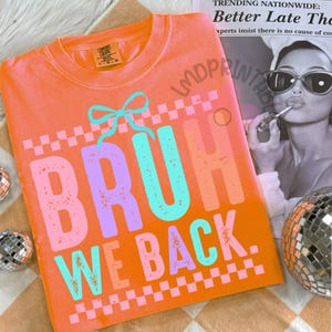Puede incluir: Camiseta naranja Comfort Colors con el texto "BRUH WE BACK" en una fuente retro colorida. El dise&ntilde;o incluye un lazo y patrones de cuadros. La camiseta se muestra sobre una superficie estampada con bolas de discoteca.