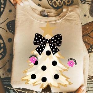 May include: A beige sweatshirt featuring a Christmas tree design. The tree has a gold star, a black and white polka dot bow, and black polka dots. Pink flowers and the text "lmdprintables" are also present.