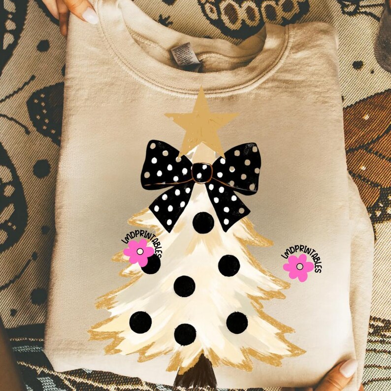 May include: A beige sweatshirt featuring a Christmas tree design. The tree has a gold star, a black and white polka dot bow, and black polka dots. Pink flowers and the text "lmdprintables" are also present.