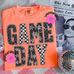 May include: Orange t-shirt with the words "GAME DAY" in a distressed, bold font. The letters feature a mix of checkered and star patterns. The shirt is a Comfort Colors brand.