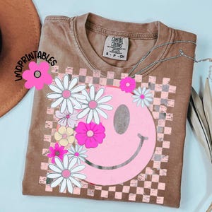 Op de afbeelding: Bruin t-shirt met een roze smiley-gezicht ontwerp, witte en roze madeliefjes en een geblokte achtergrond. Het shirt heeft de tekst "IMDPRINTABLES" en "COMFORT COLORS". Een zilveren ketting is over het shirt gedrapeerd.