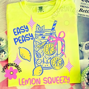 May include: A bright yellow t-shirt with a blue and pink graphic. The design shows a mason jar filled with lemons and ice cubes, with the words "EASY PEASY LEMON SQUEEZY" in blue and pink. The shirt has a round neck and short sleeves.
