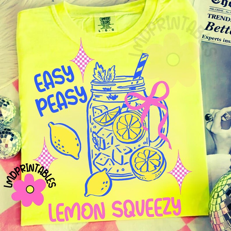 May include: A bright yellow t-shirt with a blue and pink graphic. The design shows a mason jar filled with lemons and ice cubes, with the words "EASY PEASY LEMON SQUEEZY" in blue and pink. The shirt has a round neck and short sleeves.