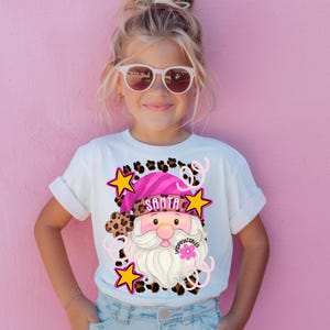 Op de afbeelding: Wit T-shirt met een kerstman-afbeelding. Het ontwerp bevat een roze hoed, witte baard, luipaardprint accenten, gele sterren en het woord "SANTA". Het T-shirt wordt gedragen door een jong persoon met een zonnebril.
