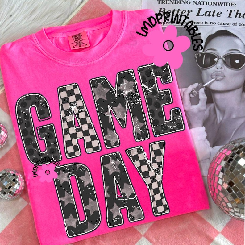 May include: A bright pink t-shirt with the words "GAME DAY" in large, distressed letters. The letters are filled with a mix of patterns, including stars, checkered squares, and leopard print. The shirt has a crew neck and short sleeves.