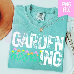 May include: A seafoam green t-shirt with the word "GARDENING" in large white letters and a row of colorful flowers. A gold necklace with a pendant is on the shirt. The shirt is folded and has a tag that says "Comfort Colors".