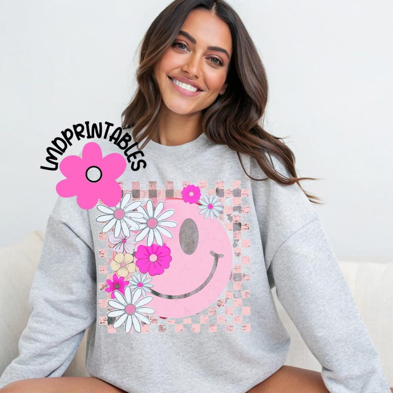 Op de afbeelding: Een lichtgrijs sweatshirt met een roze smiley-gezichtontwerp, versierd met madeliefjes en andere bloemen. Het smiley-gezicht staat tegen een roze en witte geblokte achtergrond. De tekst "LMDPRINTABLES" staat in de linkerbovenhoek.