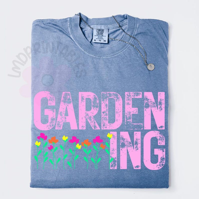May include: A blue Comfort Colors t-shirt with the word "GARDENING" in pink block letters and a row of colorful flowers. A silver necklace with a small pendant rests on the shirt.